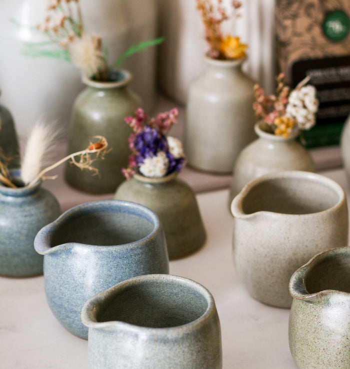 a collection of handmade jugs in various muted colours with a blurrd background of matching vases containing dried flowers 