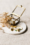 Gold hoop earrings with gold charms and green gemstones on a white coaster surface with a small glass jewellery box and dried flowers.