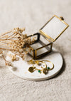 Gold hoop earrings with green stones on a speckled coaster with a glass jewellery box and dried flowers.