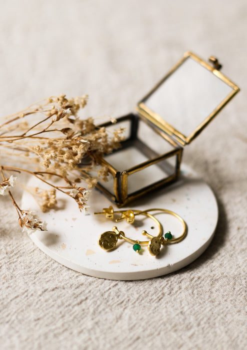 Gold hoop earrings with green stones on a speckled coaster with a glass jewellery box and dried flowers.