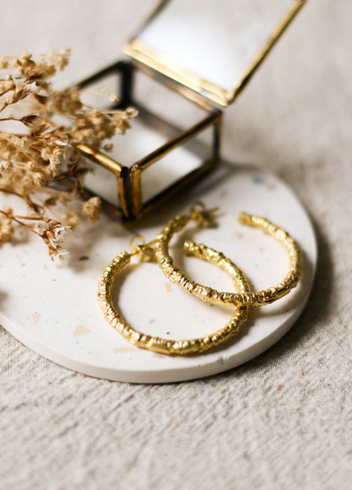 Gold hoop earrings on a white coaster with dried flowers and a gold jewellery box in the background.