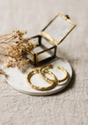 Gold hoop earrings on a white coaster with dried flowers and a small glass and brass box.