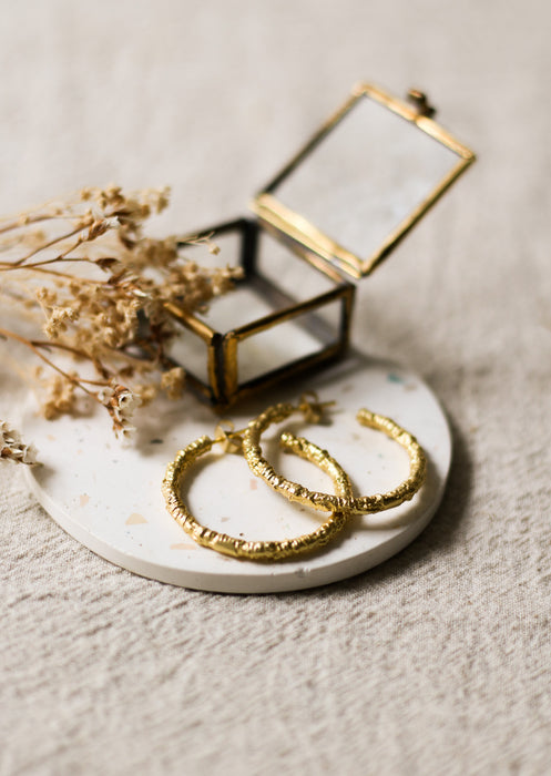 Gold hoop earrings on a white coaster with dried flowers and a small glass and brass box.