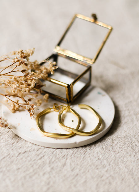 Gold hoop earrings on a white speckled coaster with a small glass jewellery box and dried flowers.