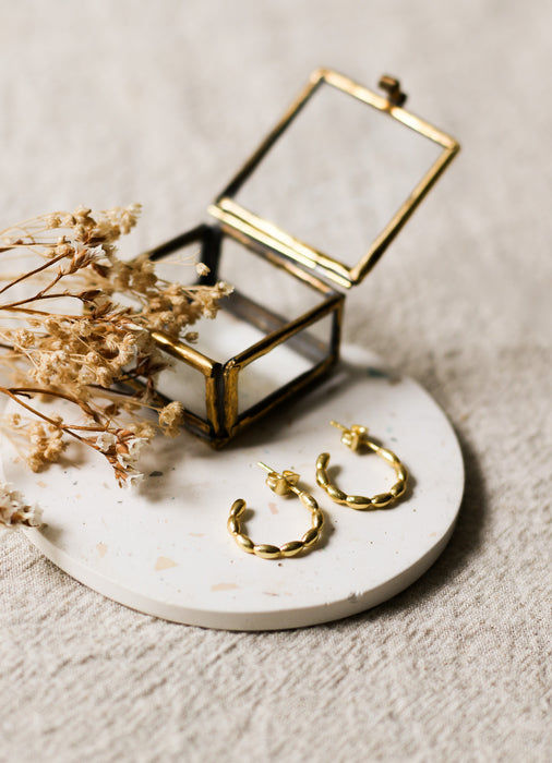 Gold hoop earrings on a white speckled coaster with a brass jewellery box and dried flowers.
