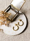 Gold hoop earrings on a white speckled coaster with a brass jewellery box and dried flowers.