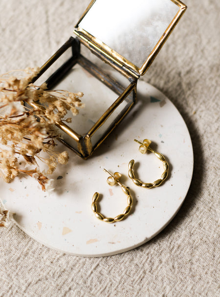 Gold hoop earrings on a white speckled coaster with a brass jewellery box and dried flowers.