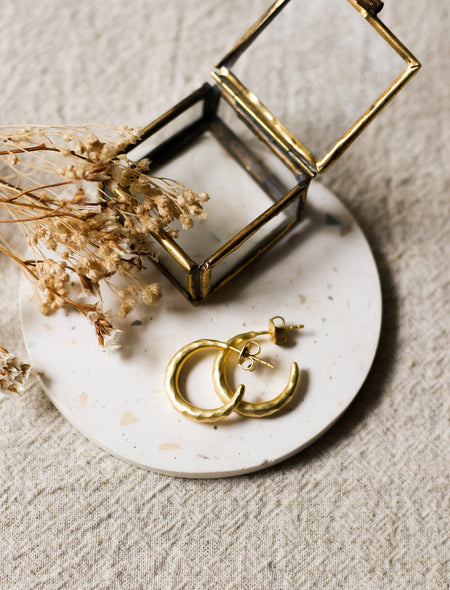 Gold earrings on a white speckled coaster with dried flowers and a gold and glass jewellery box on a beige linen surface