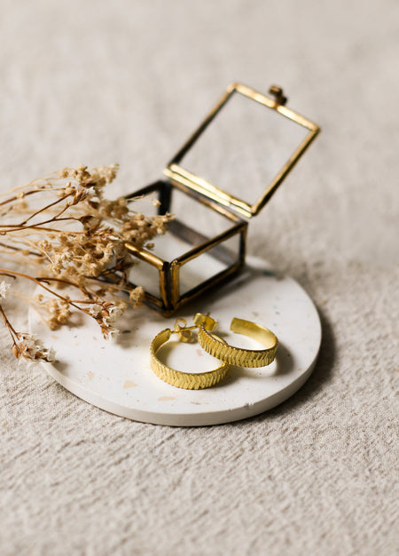 Gold hoop earrings on a white speckled coaster with dried flowers and a small glass box on a beige linen background