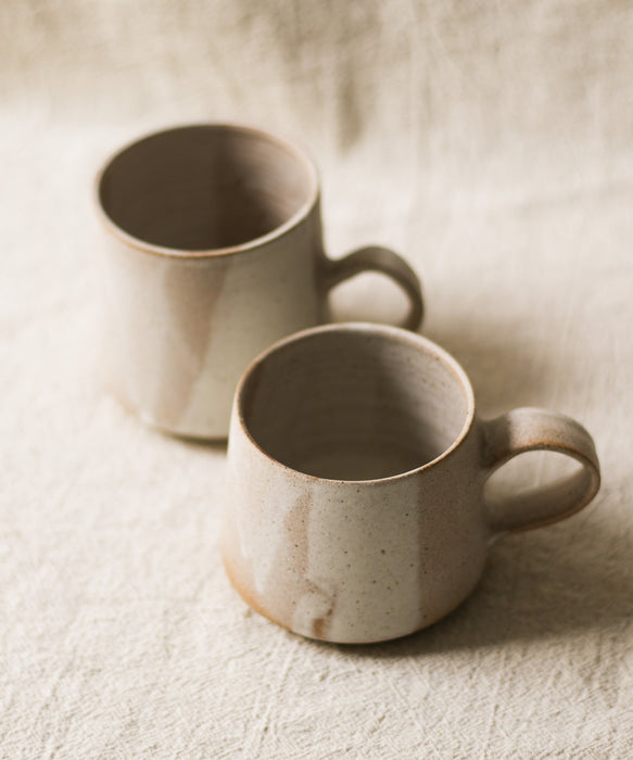 Two beige ceramic mugs on a textured surface with a neutral linen background