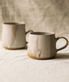 Two beige ceramic mugs on a textured surface with a neutral linen background