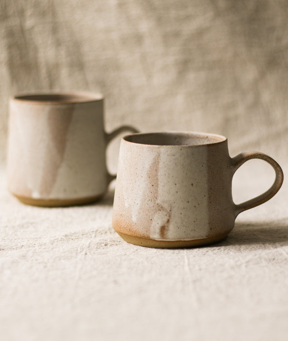 Two beige ceramic mugs on a textured surface with a neutral linen background