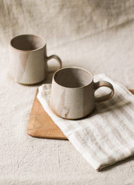 Two beige ceramic mugs on a wooden board with a white striped tea towel underneath, on a textured beige linen surface.
