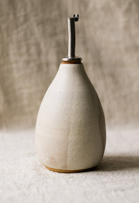 Beige ceramic oil dispenser with metal nozzle on a beige linen surface