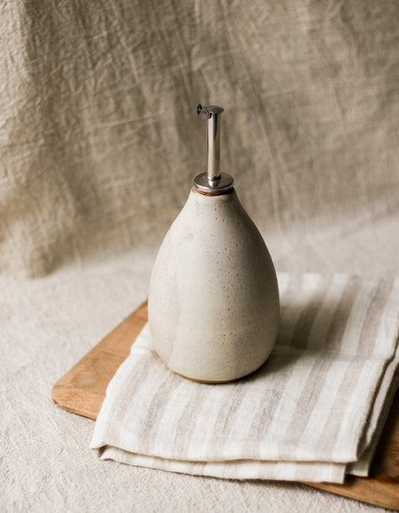 White ceramic oil dispenser on a wooden board with a striped tea towel against a beige linen background