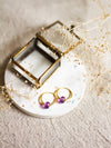 Gold hoop earrings with amethyst beads on a white dish with a brass jewellery box and dried flowers on a linen background