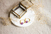 Gold hoop earrings with amethyst beads on a white dish, brass and glass jewellery box ,surrounded by dried flowers on a beige linen surface.