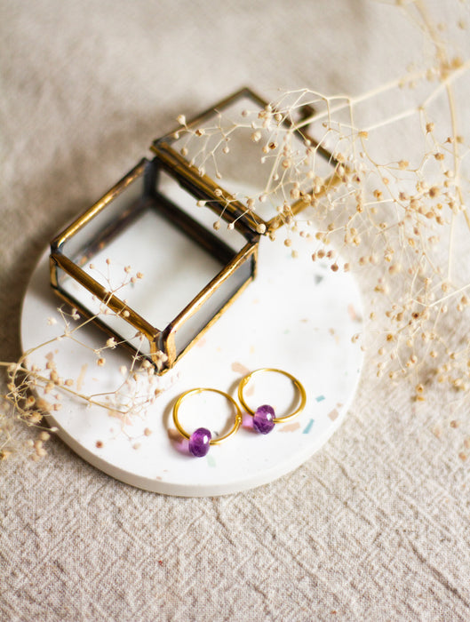 Gold hoop earrings with amethyst beads on a white dish with a brass jewellery box and dried flowers on a linen background