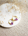 Gold hoop earrings with amethyst gemstones on a white plate with dried flowers.