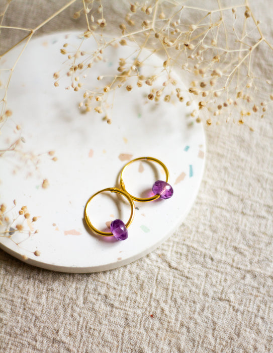 Gold hoop earrings with amethyst gemstones on a white plate with dried flowers.