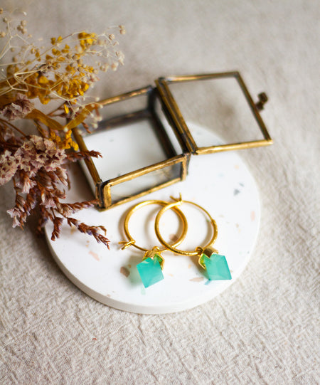Gold hoop earrings with aqua stones on a white plate with dried flowers and a brass jewellery box.