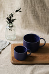 Two blue mugs on a wooden tray with a vase of greenery in the background.