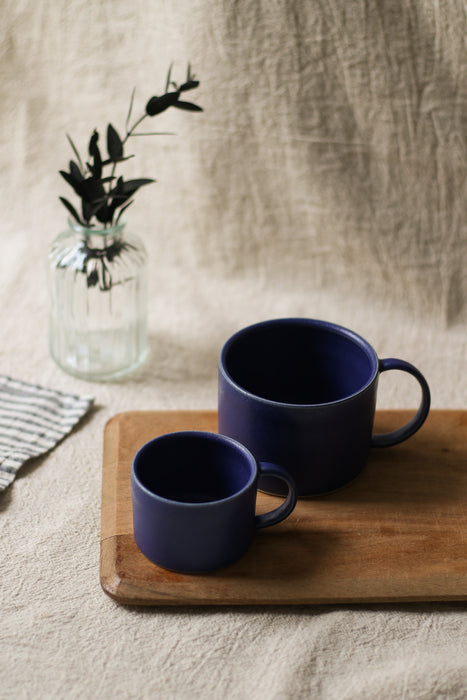 Two blue mugs on a wooden tray with a vase of greenery in the background.