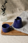 a blue ceramic cup and jug on a wooden board with a striped tea towel and leaves in a vase in the background.