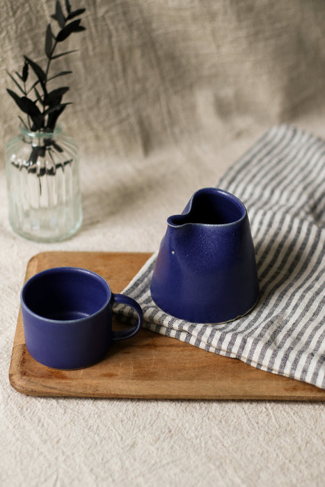 a blue ceramic cup and jug on a wooden board with a striped tea towel and leaves in a vase in the background.