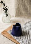 Navy blue ceramic pitcher on a wooden board with a striped cloth and vase of leaves in the background