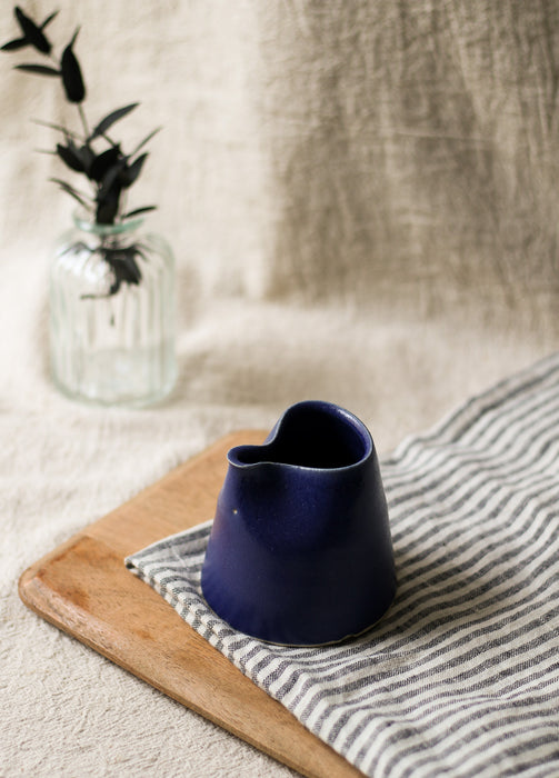 Navy blue ceramic pitcher on a wooden board with a striped cloth and vase of leaves in the background