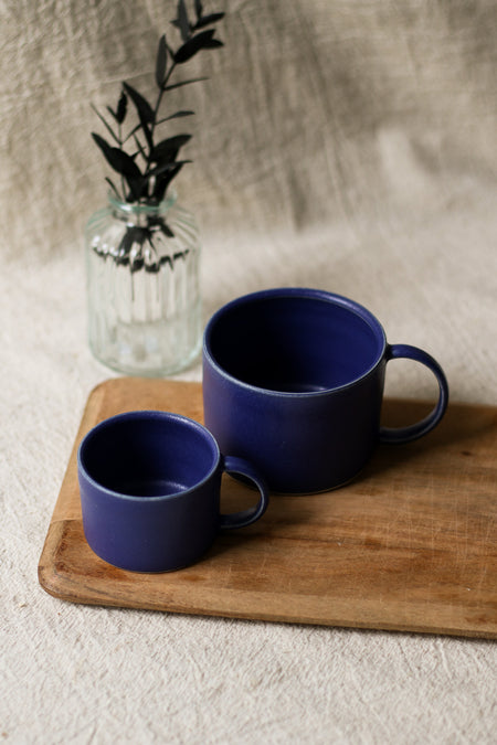 Two blue mugs on a wooden tray with a glass vase containing plants in the background.