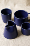 Three blue ceramic mugs and a jug on a textured linen surface