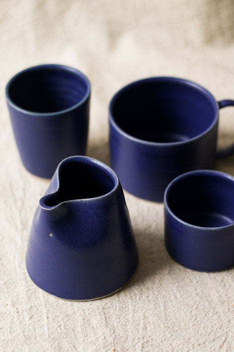 Three blue ceramic mugs and a jug on a textured linen surface