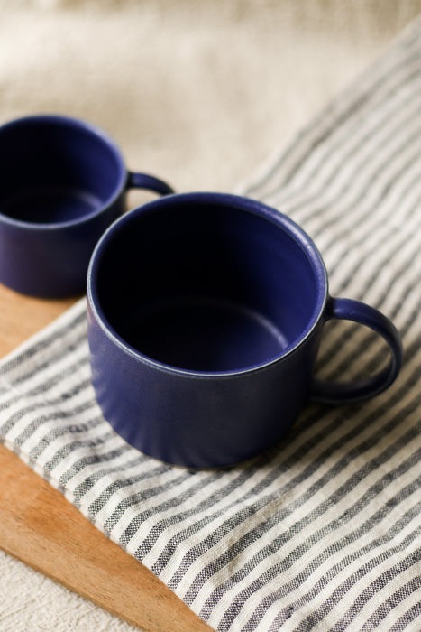 Two blue mugs on a striped tea towel and a wooden bread board