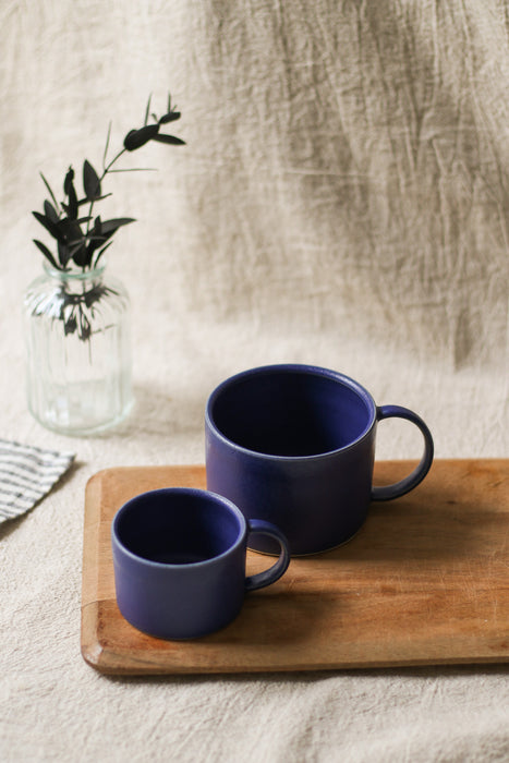 Two blue mugs on a wooden board with a vase of greenery in the background.