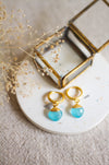 Gold hoop earrings with aqua drops on a white plate with dried flowers and a gold box.