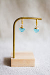 Gold hoop earrings with aqua gemstones on a wooden stand against a neutral background