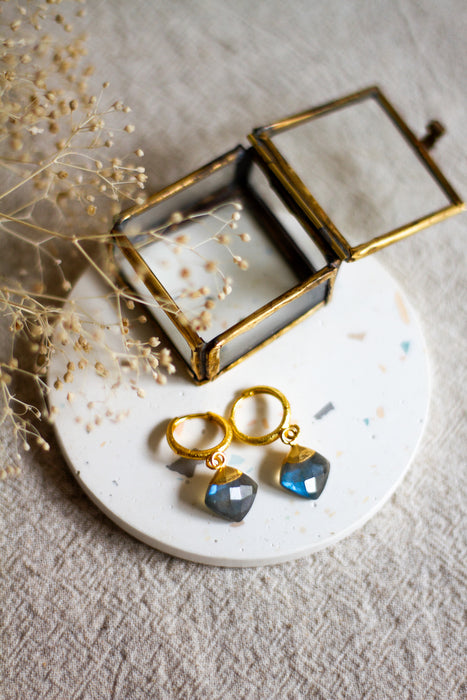 Gold hoop earrings with labradorite drops on a white plate with a gold box and dried flowers.