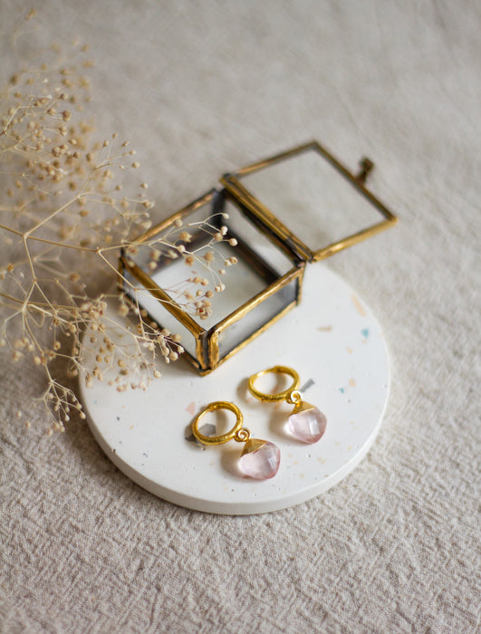 Gold earrings with rose quartz stones on a white plate with a gold jewellery box and dried flowers.
