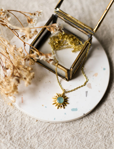Gold necklace with a gold sun shaped pendant set with a gemstone on a white coaster with dried flowers