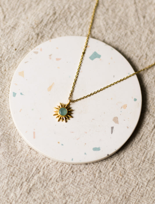 Gold necklace with a sun pendant on a round, speckled coaster on a beige linen surface