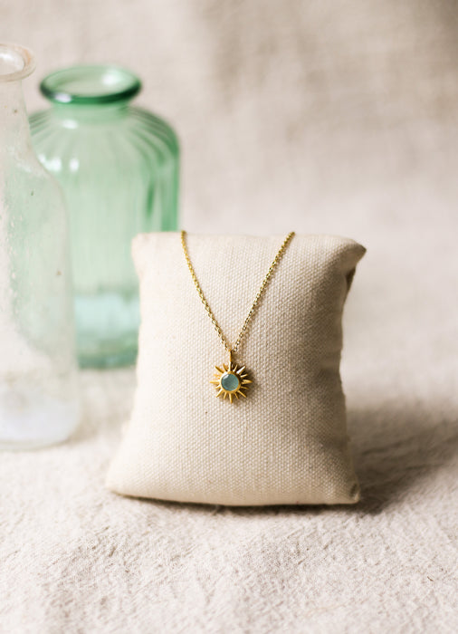 Gold necklace with a sun pendant on a beige pillow with a blurred green vase in the background.