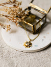 Gold necklace with a sun pendant on a white surface with dried flowers.