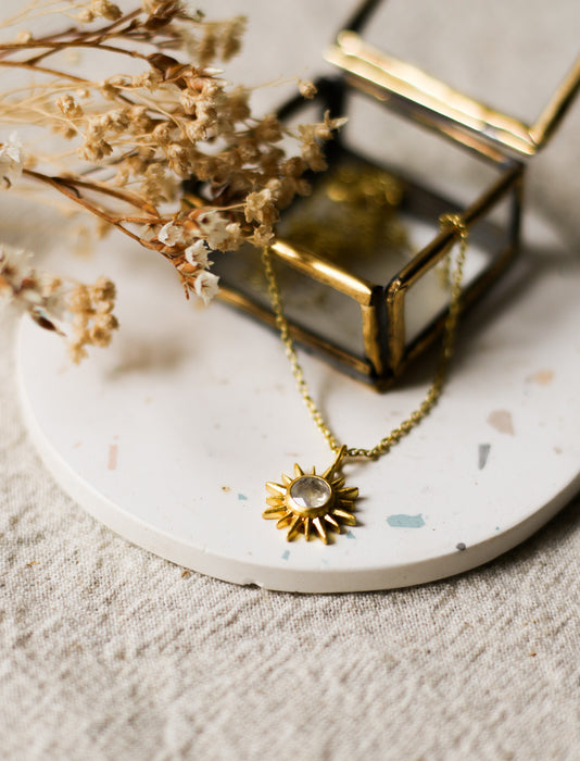 Gold necklace with a sun pendant on a white surface with dried flowers.