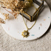 Gold necklace with a gold sun shaped pendant set with a gemstone on a white coaster with dried flowers