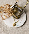 Gold necklace with a gold sun shaped pendant set with a gemstone on a white coaster with dried flowers
