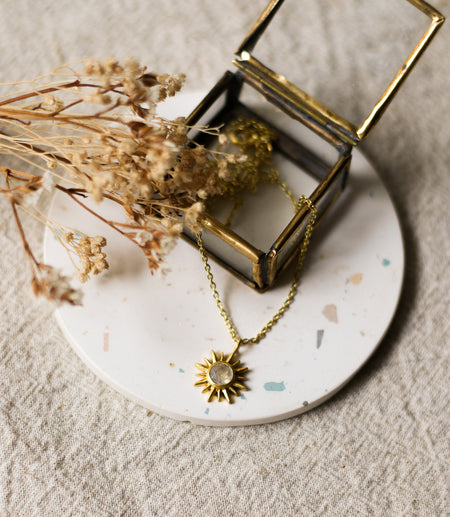 Gold necklace with a gold sun shaped pendant set with a gemstone on a white coaster with dried flowers