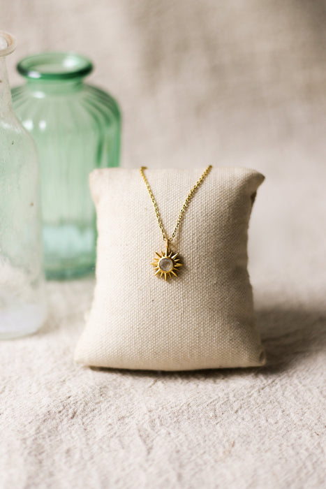Gold necklace with a sun pendant on a beige pillow with a blurred green vase in the background.