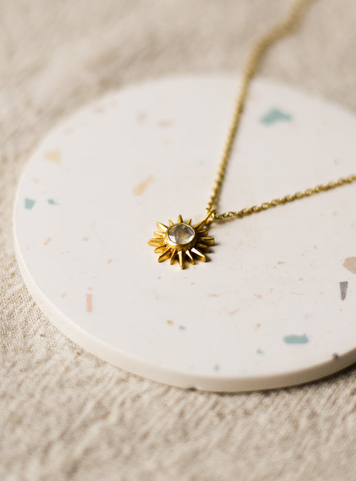 Gold necklace with a sun pendant on a round, speckled coaster.